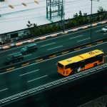 Ônibus urbano amarelo circula por uma avenida movimentada, visto de cima, entre outros veículos.