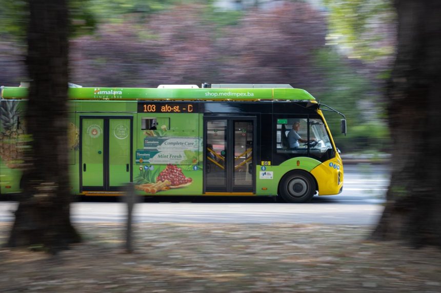 Ônibus elétrico verde circula por uma avenida movimentada de Sarajevo, entre carros e edifícios urbanos.